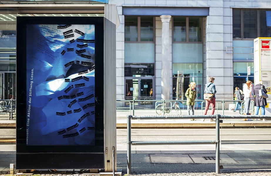 a train station in a city with a large building in the background, waiting passengers, and a billboard with a poster on which in bluish tint is a crumpled sheet and a poem.